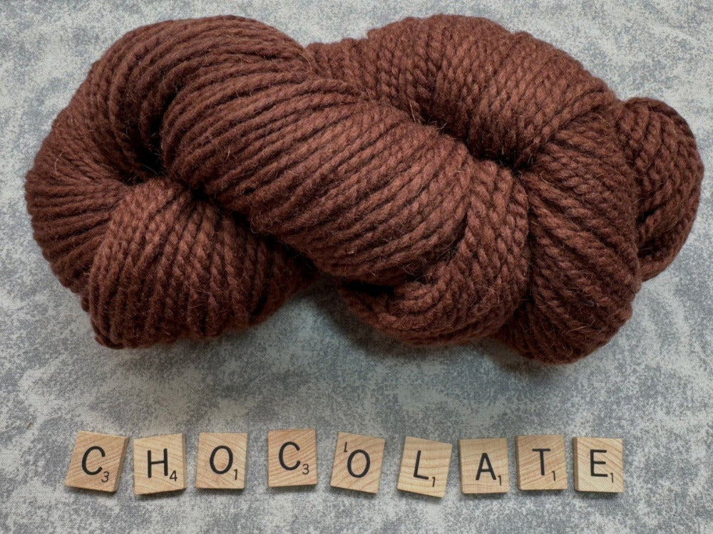 Brown skein of yarn with 'CHOCOLATE' spelled out using wooden tiles on a textured gray background